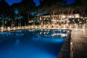 A nighttime pool area with a wooden deck, trees, and a building in the background. - Hotel Fontermosa (Orbetello)