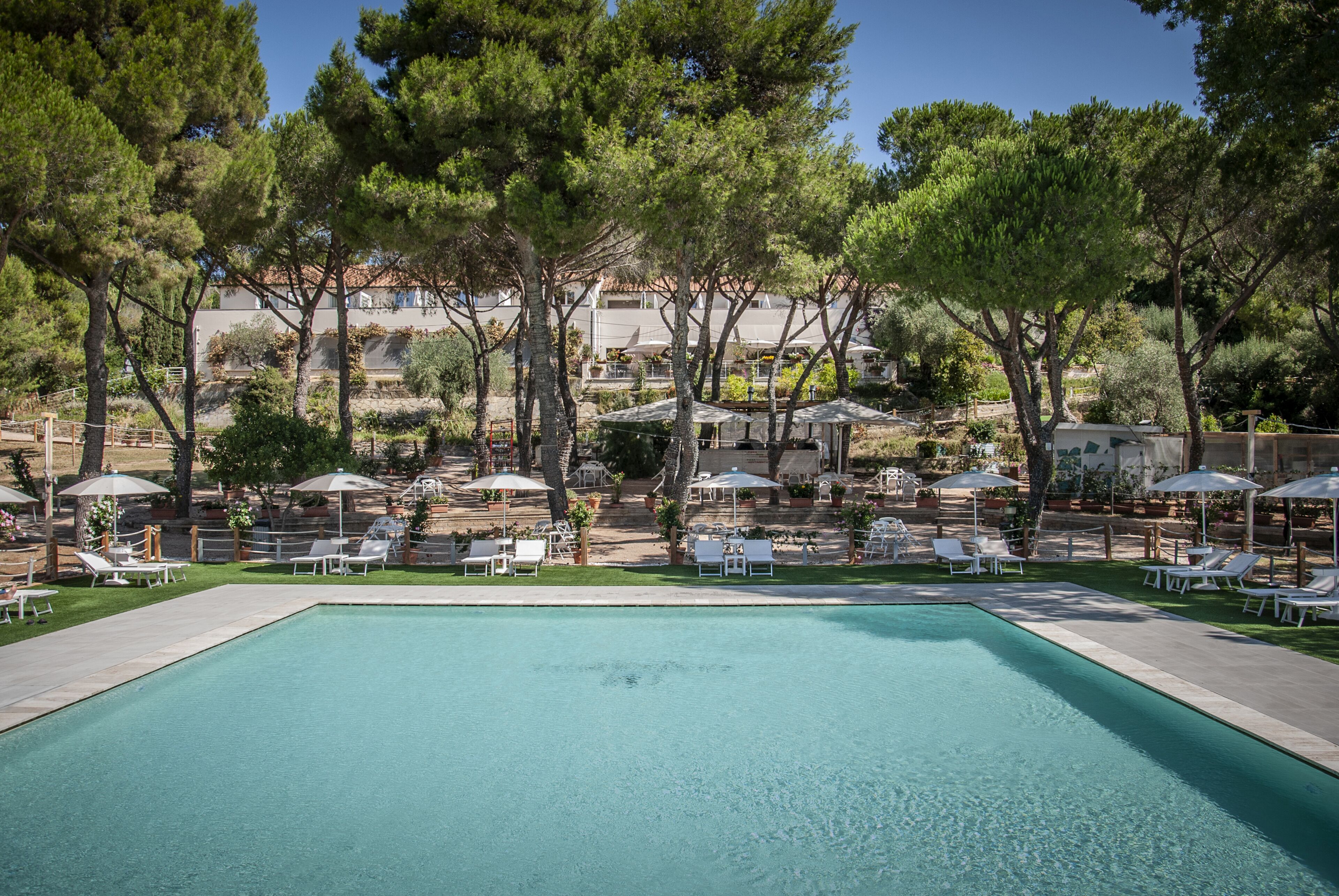 Una piscina con lettini e ombrelloni, circondata da alberi e un edificio sullo sfondo.