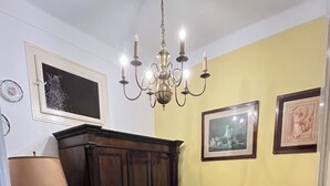 A room with a wooden floor, a dark wooden wardrobe, a white sofa, a small bedside table with a lamp, and a chandelier.