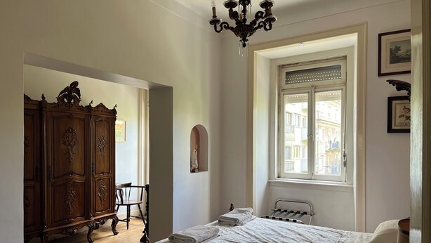 A bedroom with a bed, a wooden wardrobe, a chandelier, a window with a view of buildings, and a door.