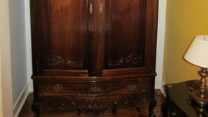 A large wooden wardrobe with ornate carvings and brass handles, positioned in a room with a wooden floor and a painting on the wall.