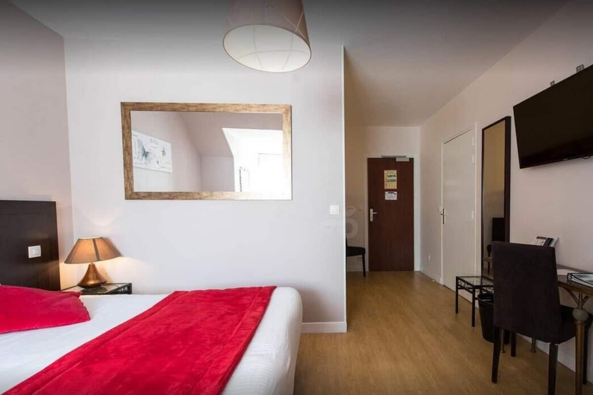 A hotel room with a bed, a desk, a chair, a TV, a mirror, and a door leading to another room.