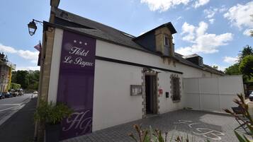 Un hotel llamado 'Hotel Le Bayeux' con un letrero púrpura, entrada pavimentada y un pequeño jardín.