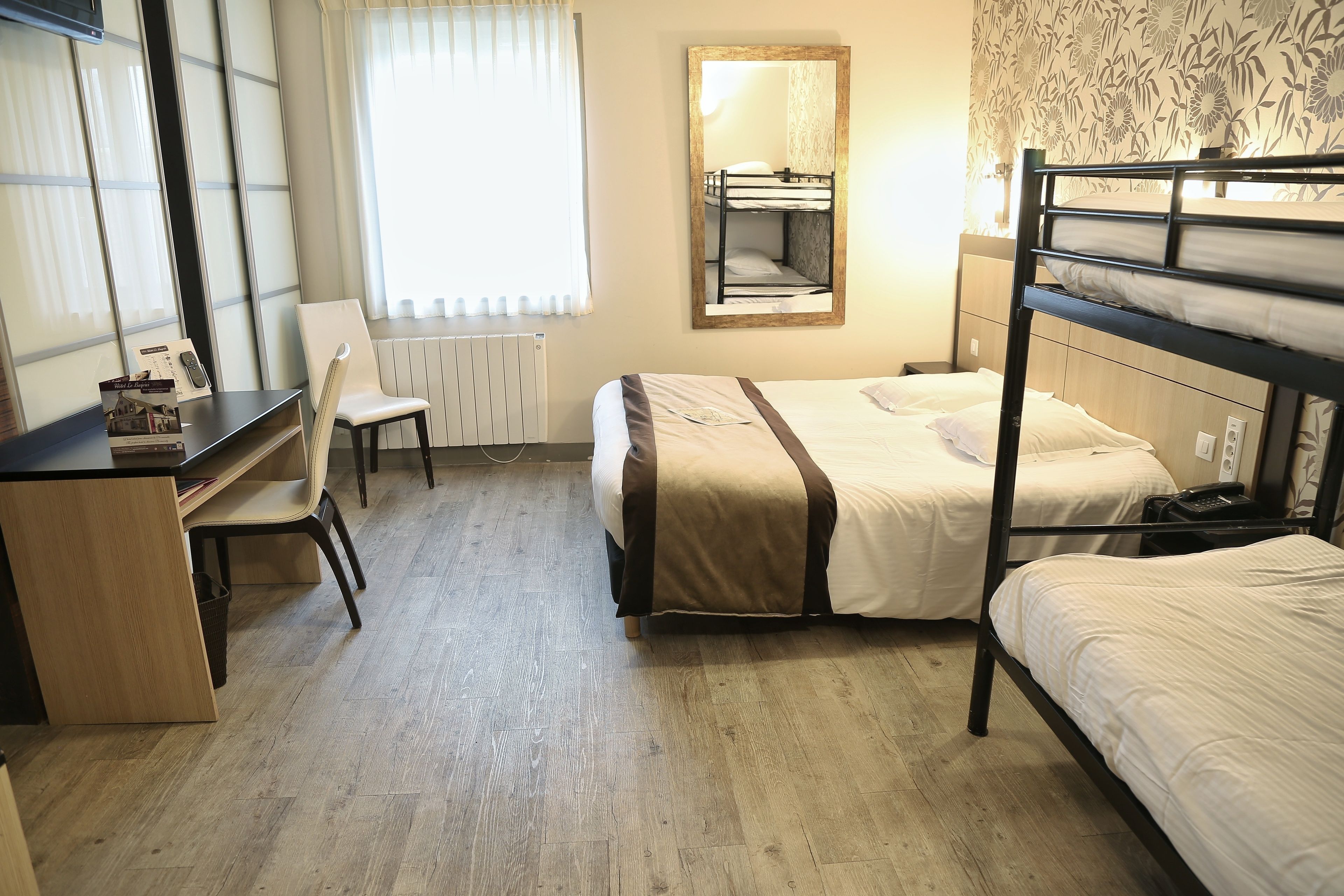 A hotel room with a bunk bed, a desk with a chair, a radiator, and a window with curtains.