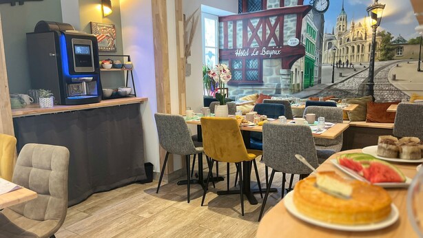 A restaurant with a view of a cityscape, including a bridge and a church. There are tables with chairs, a coffee machine, and a counter with a menu. - Hotel Le Bayeux (Bayeux)