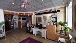 A reception area with a wooden desk, a computer, and a bulletin board. Flags are hanging from the ceiling, and there is a plant on the desk. - Hotel Le Bayeux (Bayeux)
