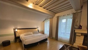 A bedroom with a bed, a nightstand, a wooden floor, a window with curtains, and a fireplace. - Hotel Le Bayeux (Bayeux)