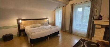 Un dormitorio con una cama, una mesita de noche, suelo de madera, una ventana con cortinas y una chimenea.