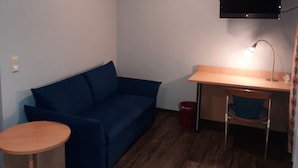 A small room with a blue sofa, a wooden desk, a chair, a small table, and a mounted TV.