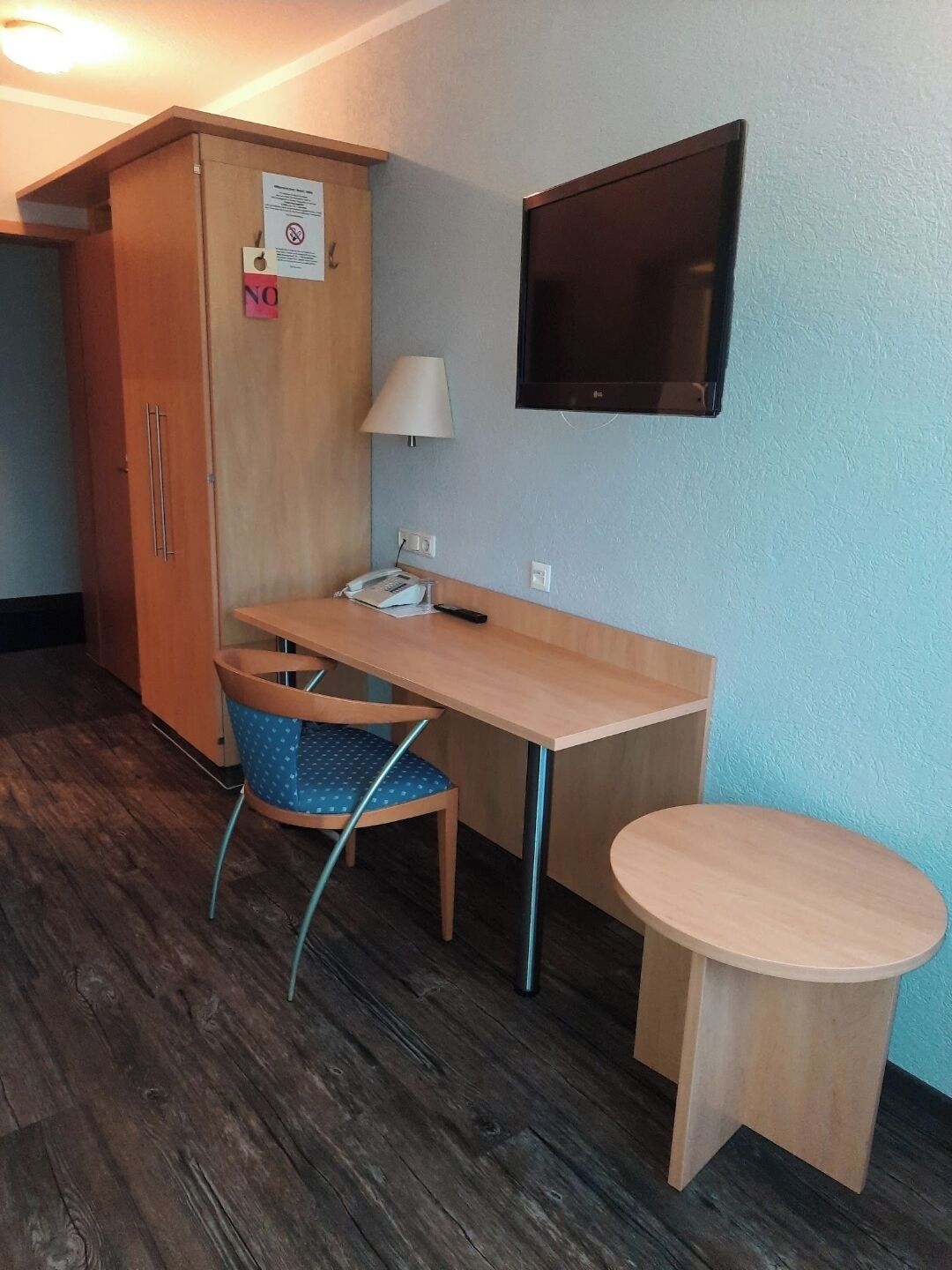 Ein gemütliches Zimmer mit Holzschreibtisch, einem Sessel, einem kleinen Tisch, einem an der Wand befestigten Fernseher und einem Kleiderschrank.