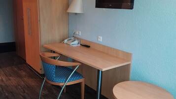 Ein gemütliches Zimmer mit Holzschreibtisch, einem Sessel, einem kleinen Tisch, einem an der Wand befestigten Fernseher und einem Kleiderschrank.
