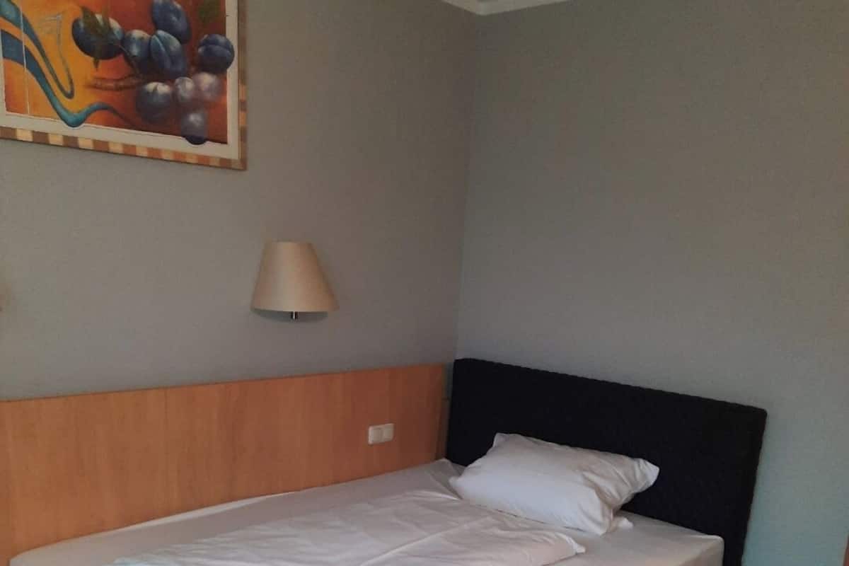 Un lit simple avec une tête de lit en bois et une literie blanche, dans une pièce dont le sol est en bois et où l’on peut voir un tableau accroché au mur.
