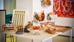 Uma mesa com diversos alimentos, incluindo doces e biscoitos, disposta em um quarto com um quadro abstrato colorido na parede.