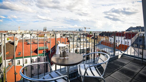 Varanda com mesa e cadeiras, vista para paisagem urbana com edifícios e guindastes.