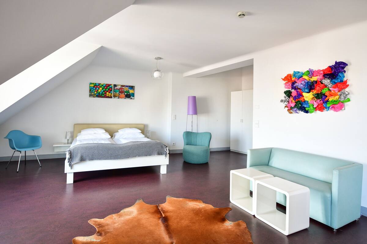 A modern bedroom with a bed, a blue chair, a teal armchair, a white side table, and a colorful wall art piece.
