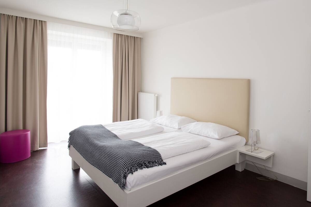 A modern hotel room with a large bed, a bedside table, a nightstand, a lamp, and a door.