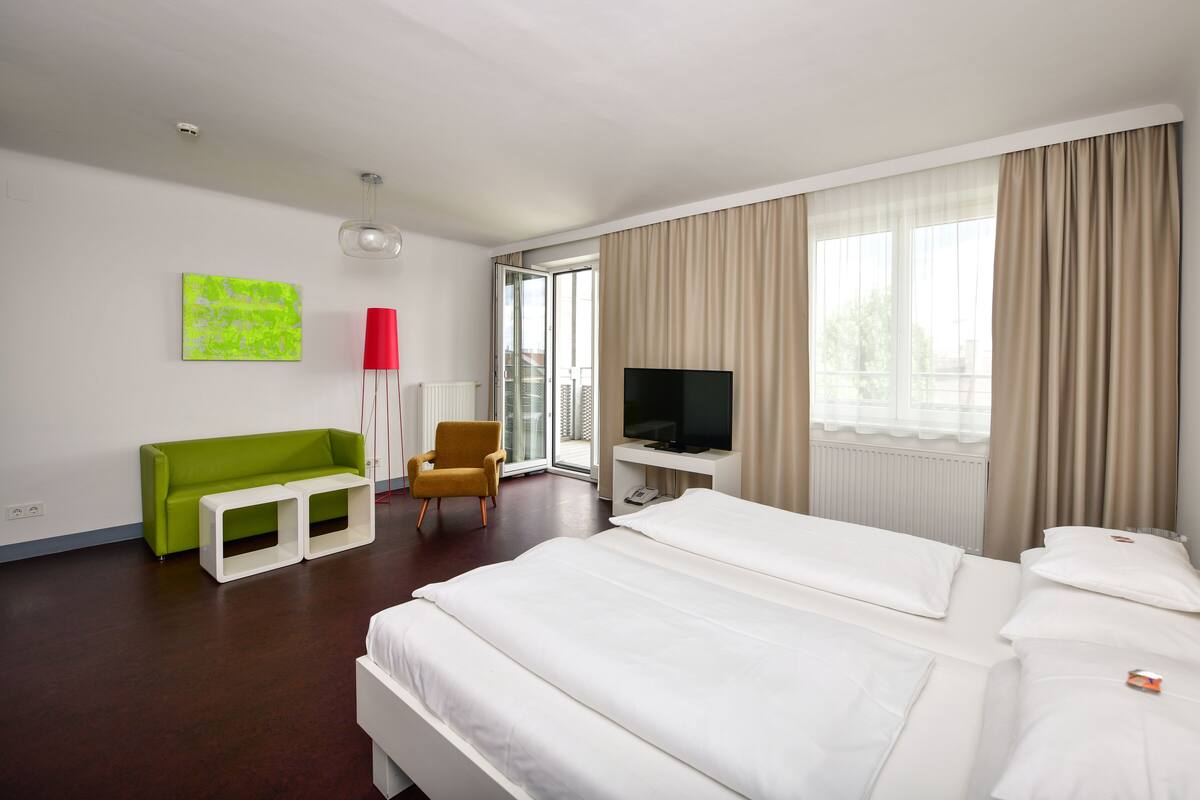 A hotel room with a bed, a desk with a TV, a green sofa, a red floor lamp, and a window with curtains.