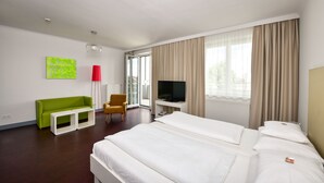 Quarto de hotel com cama, escrivaninha com TV, sofá verde, candeeiro de chão vermelho e janela com cortinas.