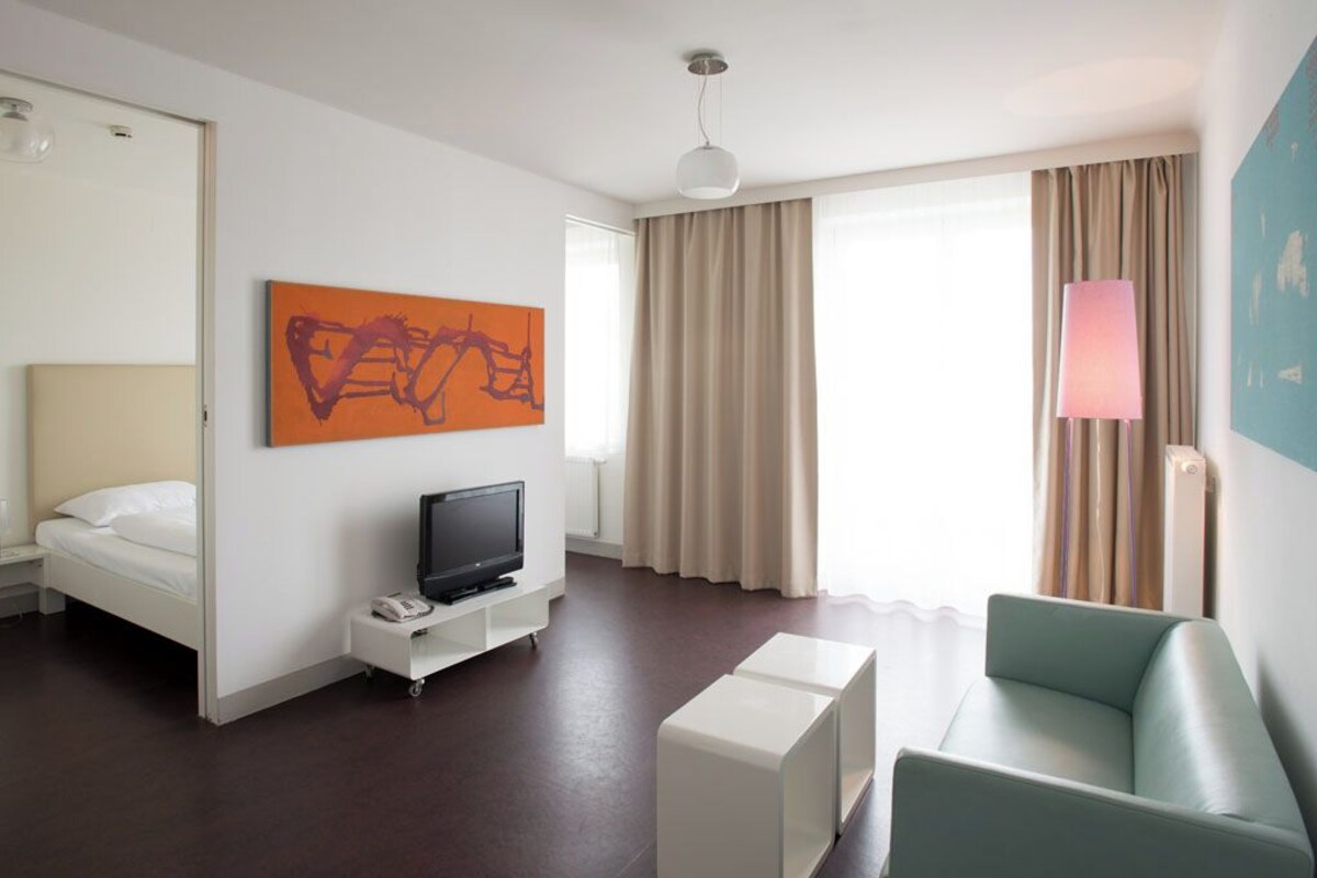 A hotel room with a bed, a TV on a stand, a desk, a chair, and a large window with curtains.