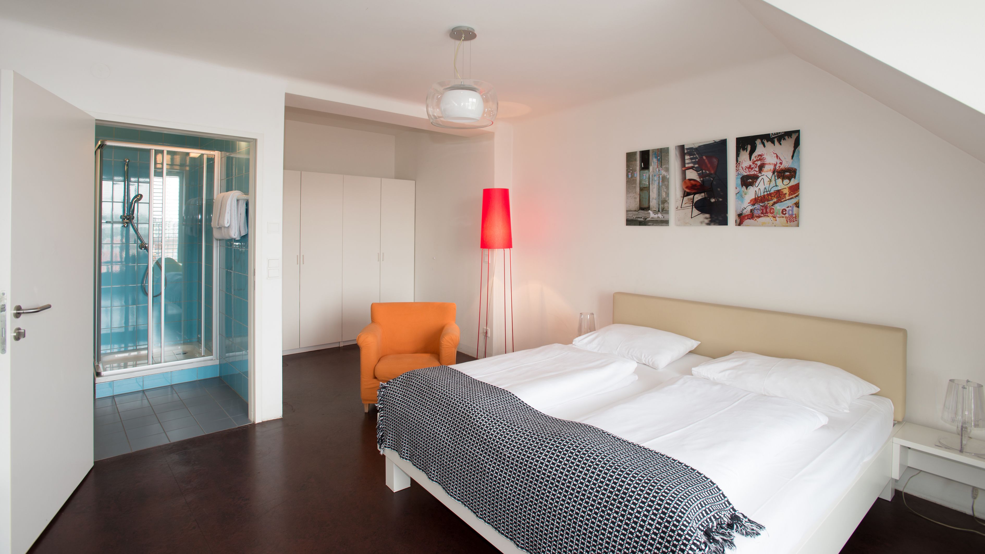 A hotel room with a bed, a chair, a red floor lamp, and a shower area.
