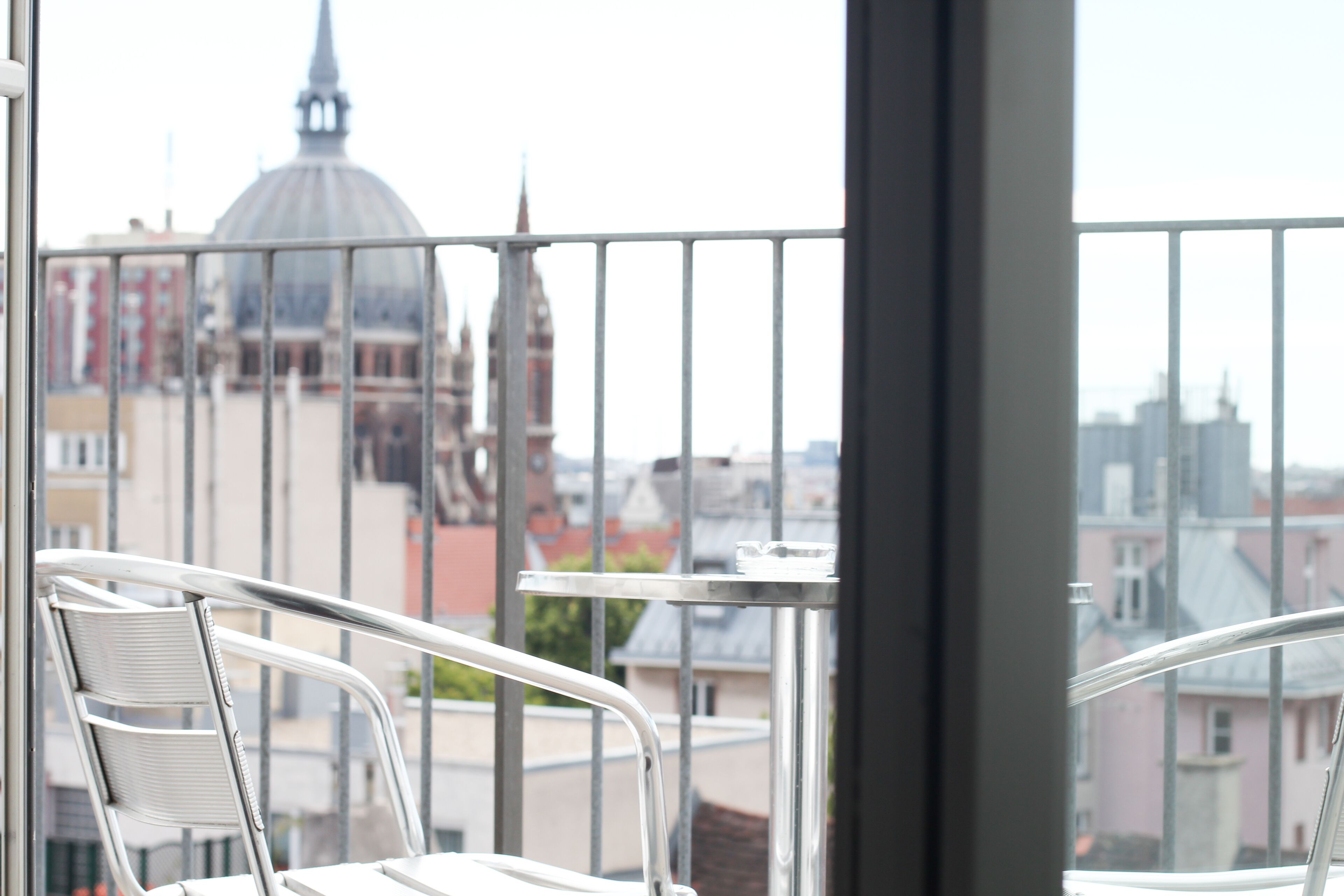 Ein Balkon mit Metallgeländern, einem weißen Stuhl und einem kleinen Tisch. Im Hintergrund ist eine Stadtansicht mit Gebäuden und einer kuppelförmigen Struktur zu sehen.