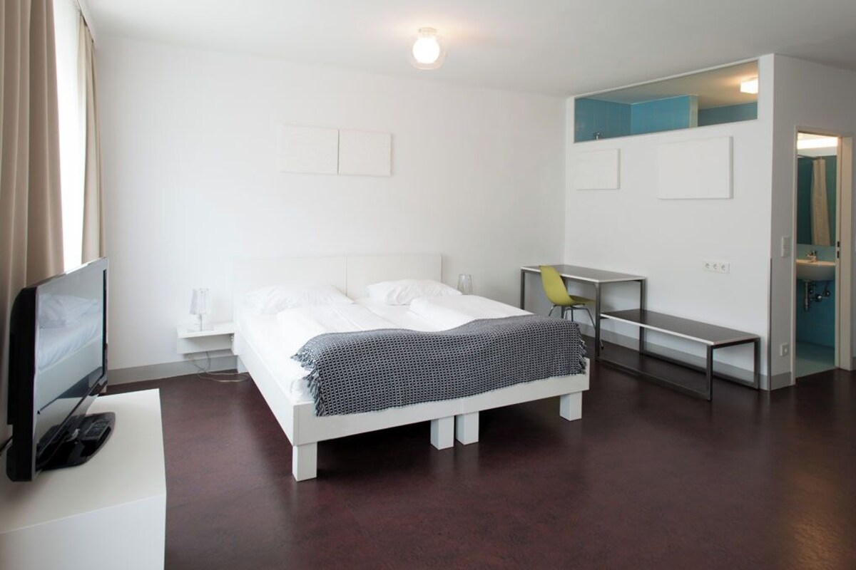 A modern hotel room with a bed, desk, chair, TV, and a bathroom visible through an open door.