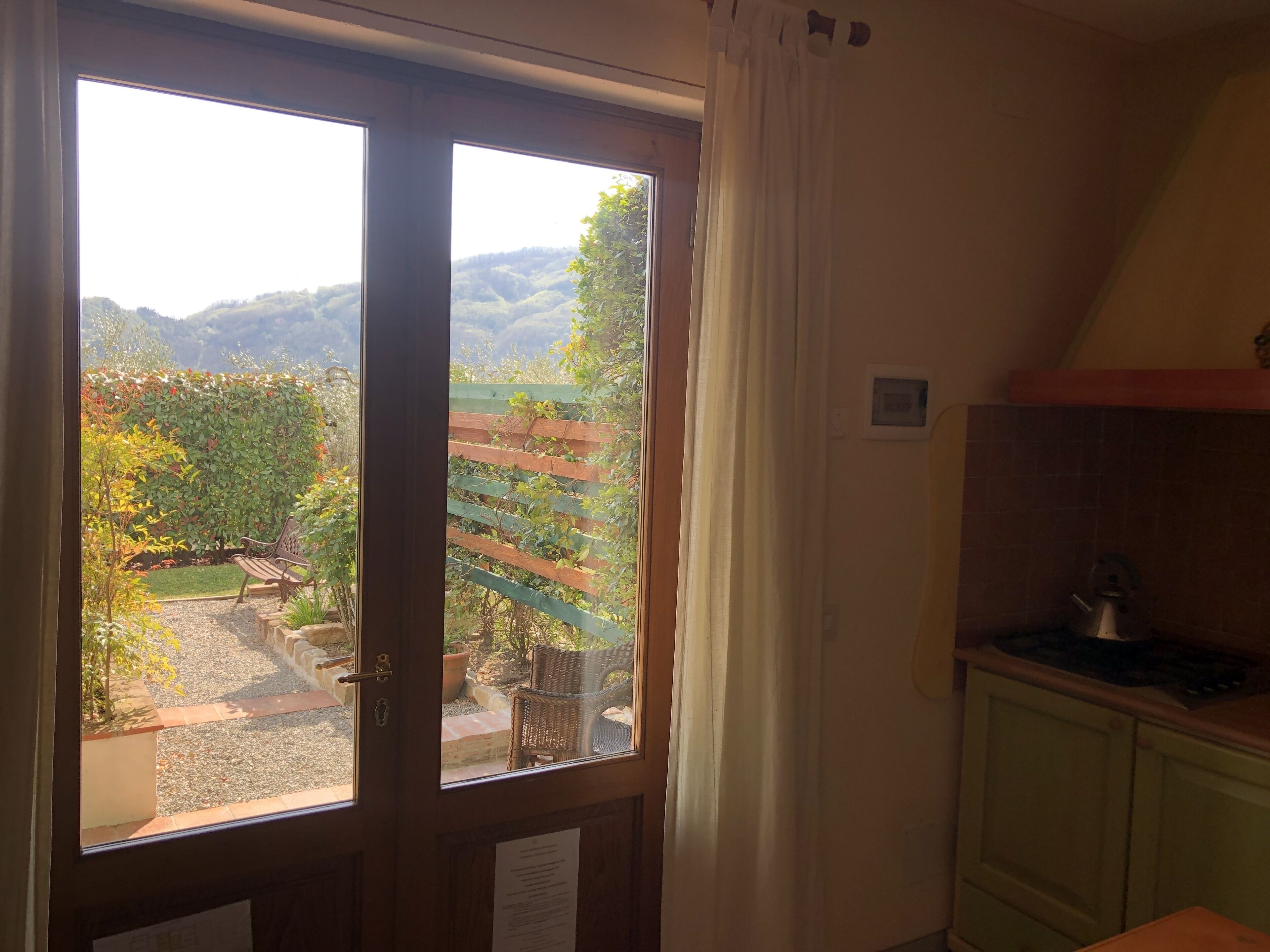 Une pièce dotée d’une grande porte-fenêtre coulissante donnant sur un jardin avec une clôture en bois, une chaise en osier et une petite table sur laquelle se trouve une bouilloire.
