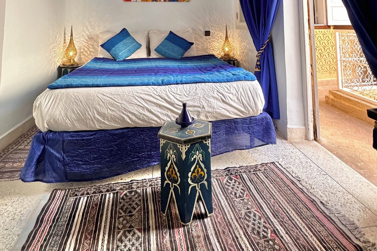 A bedroom with a bed, blue curtains, a decorative ceiling light, a colorful painting, and a patterned rug.