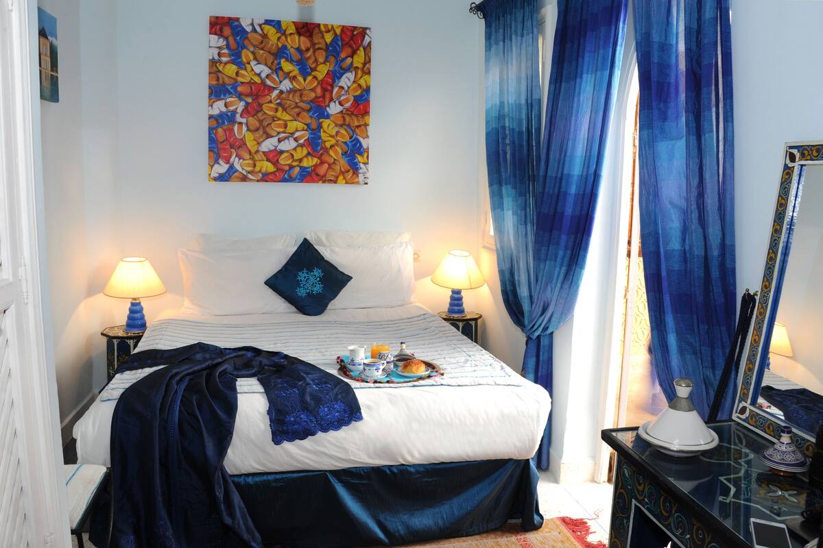 Een slaapkamer met een bed, twee bedlampen, een dienblad met ontbijtartikelen, een kleurrijk kunstwerk aan de muur en een spiegel met decoratieve lijst.