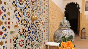 Un mur recouvert de carreaux aux motifs géométriques complexes, un pot en céramique décoratif doté d’un couvercle et une petite table sur laquelle est posé un bouquet de fleurs.