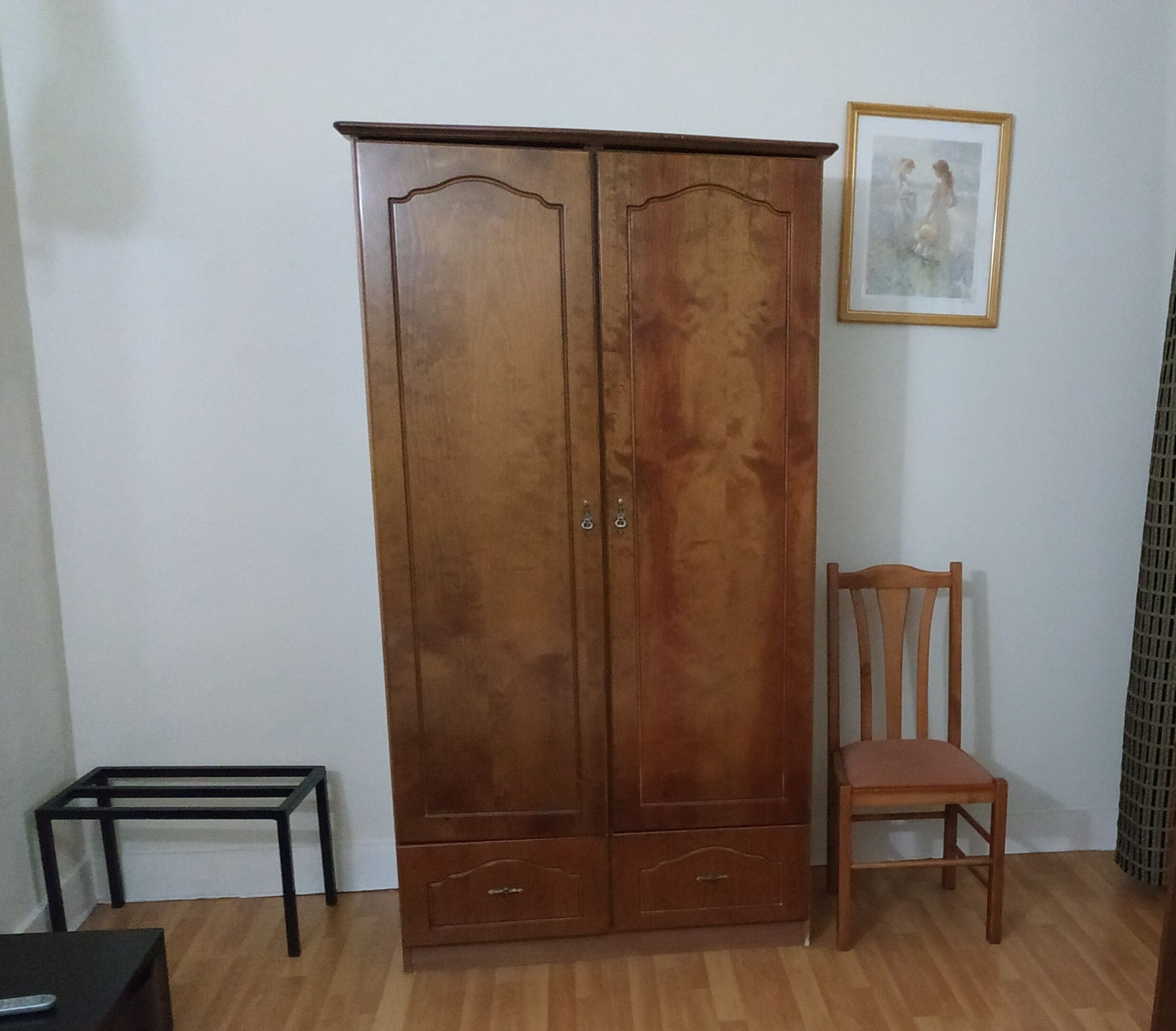 A wooden wardrobe with two doors, a small table, and a chair in a room with wooden flooring and a plain wall.