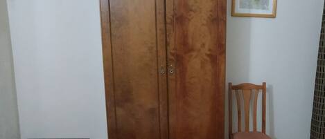 A wooden wardrobe with two doors, a small table, and a chair in a room with wooden flooring and a plain wall.