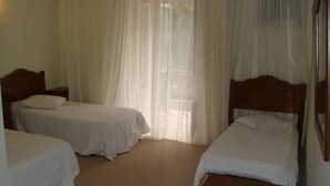 Une chambre avec deux lits simples, une tête de lit en bois, une table de chevet avec une télécommande et des rideaux légers.