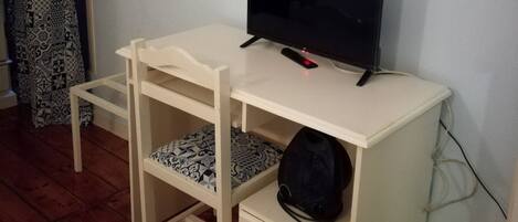 Une petite pièce avec un bureau blanc, une télévision, un rideau à motifs et un miroir au mur.