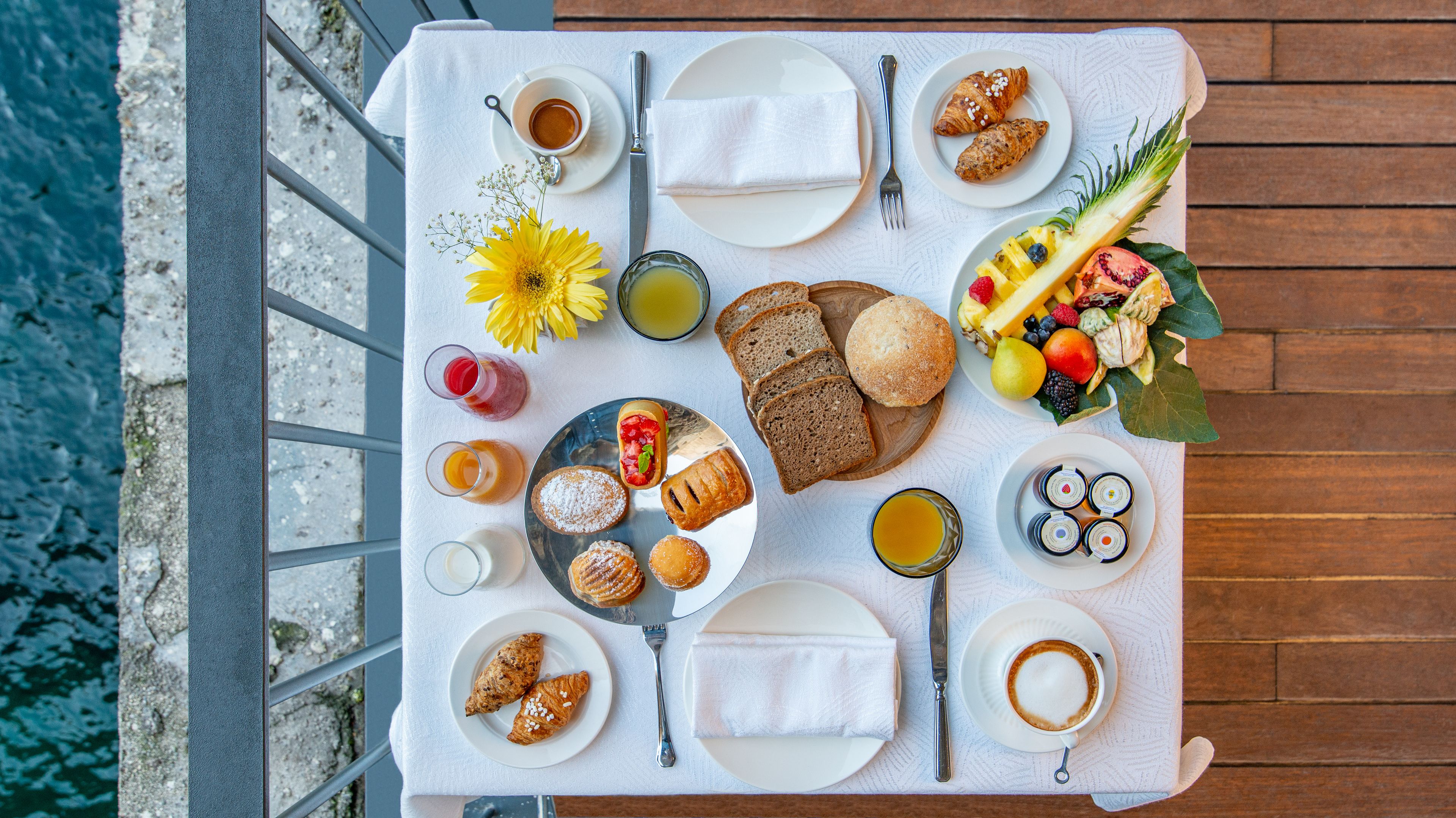 Una tavola imbandita per la colazione con croissant, pane, frutta, pasticcini e bevande, con vista sul mare sullo sfondo.