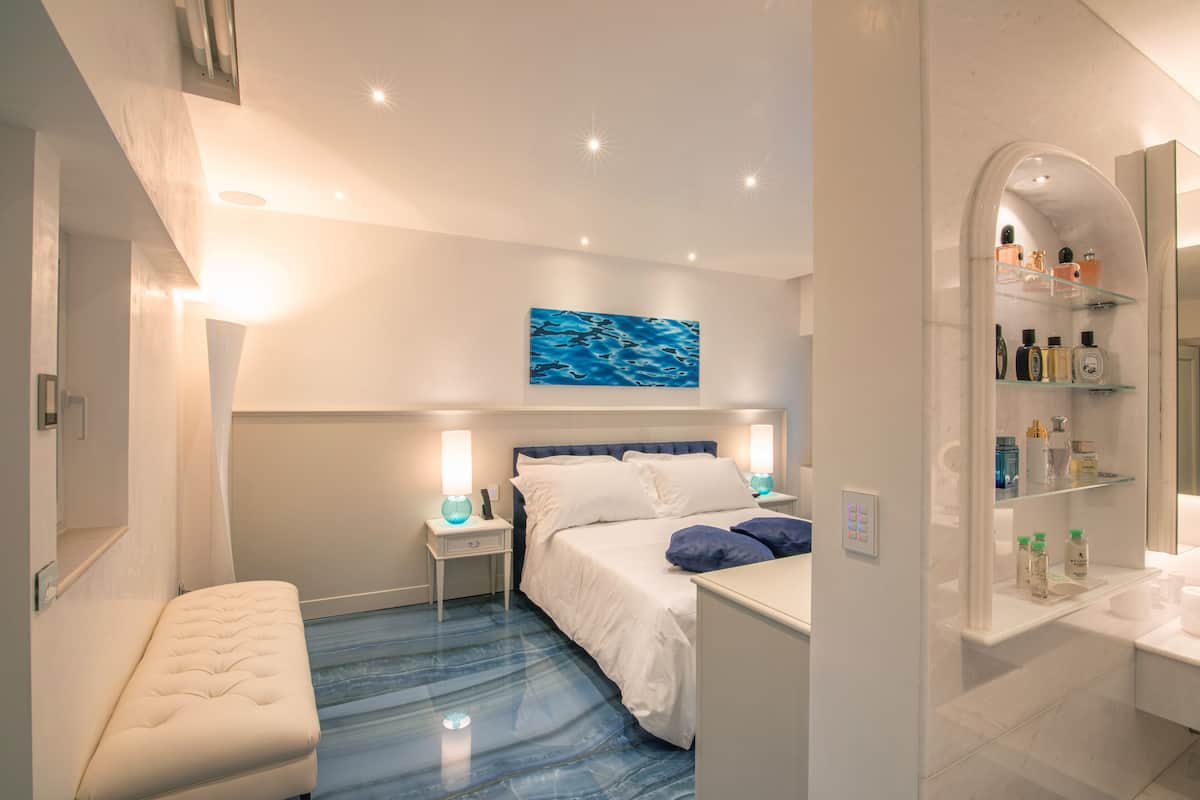 A modern bedroom with a bed, bedside tables, a bench, a blue artwork, and a bathroom with shelves.