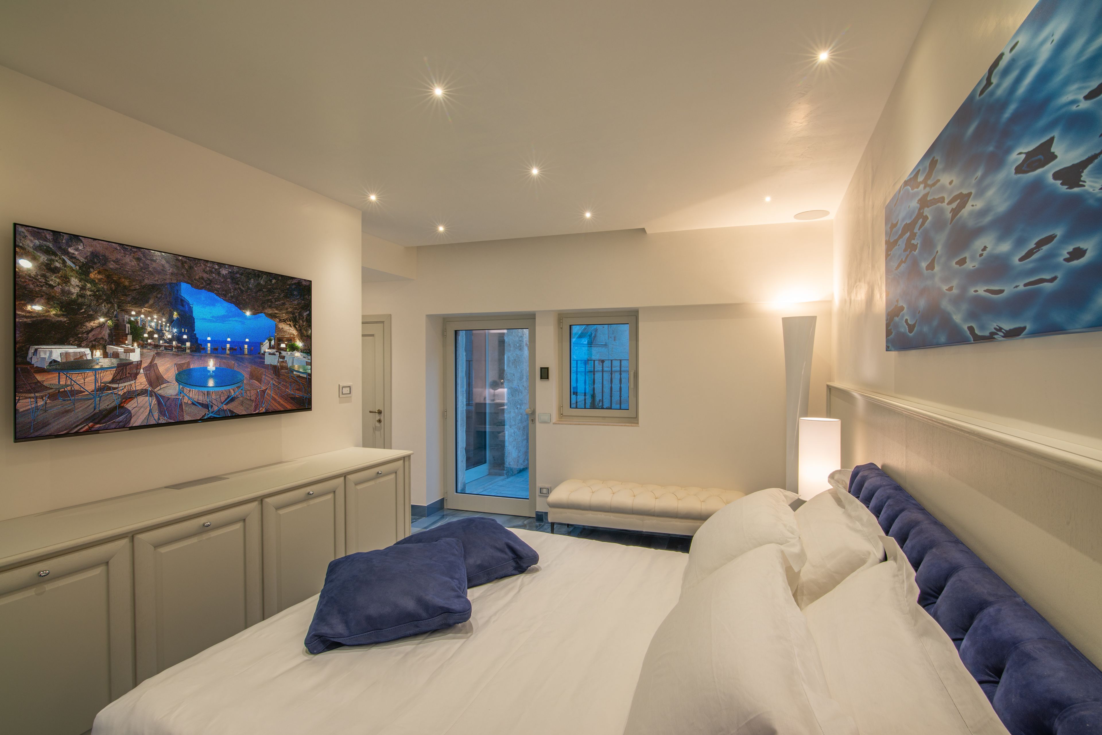 Une chambre d’hôtel moderne avec un grand lit, un téléviseur à écran plat fixé au mur, un banc et une porte donnant sur un balcon.