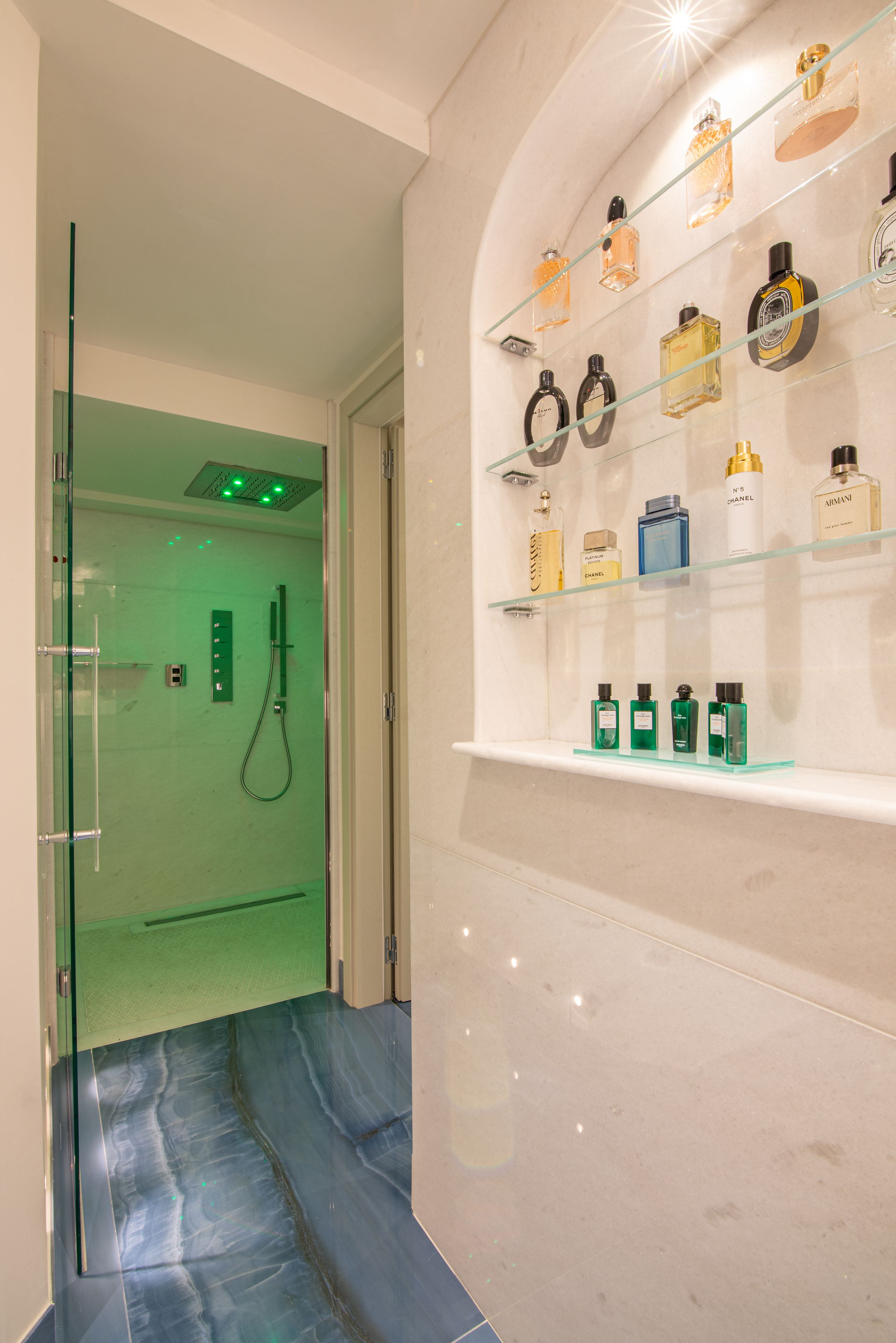 A modern bathroom with a glass shower enclosure, a marble floor, and a shelf with various toiletries.