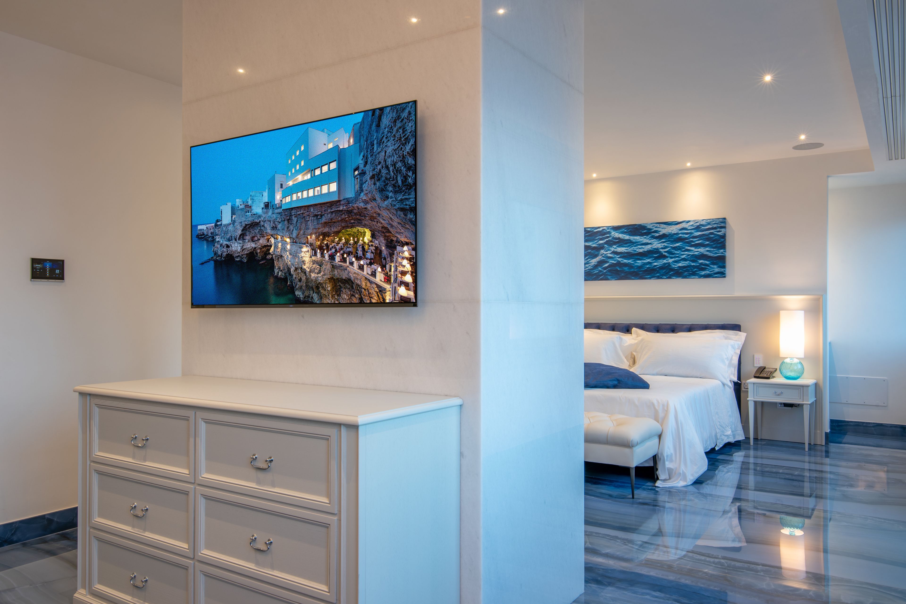 Ein modernes Hotelzimmer mit einem großen Bett, einem Nachttisch mit Lampe, einem an der Wand montierten Flachbildfernseher und einer weißen Kommode.