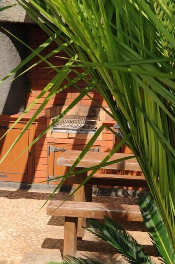 A brick barbecue grill with a wooden table in front, surrounded by greenery.