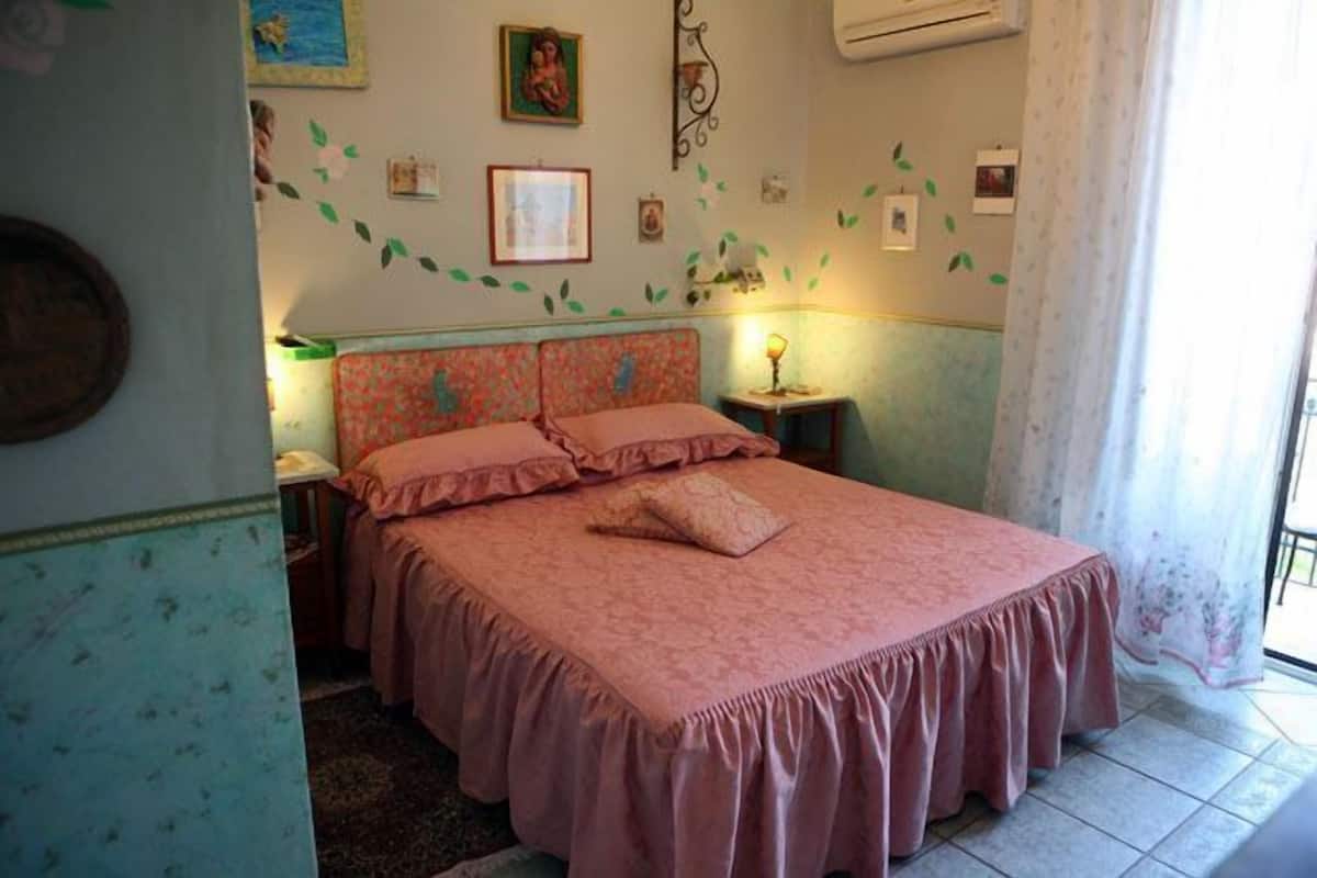 A bedroom with a bed, a nightstand, a wall-mounted lamp, and a window with curtains.