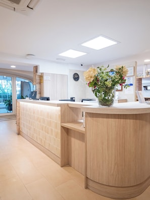 A reception desk with a vase of flowers, a clock on the wall, and a view of the outdoors. - Hotel Bersoca (Benicassim)