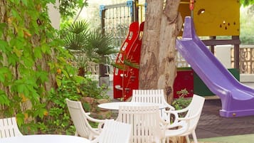An outdoor patio with white plastic chairs and tables, a tree, and a playground with a red slide and a purple slide.