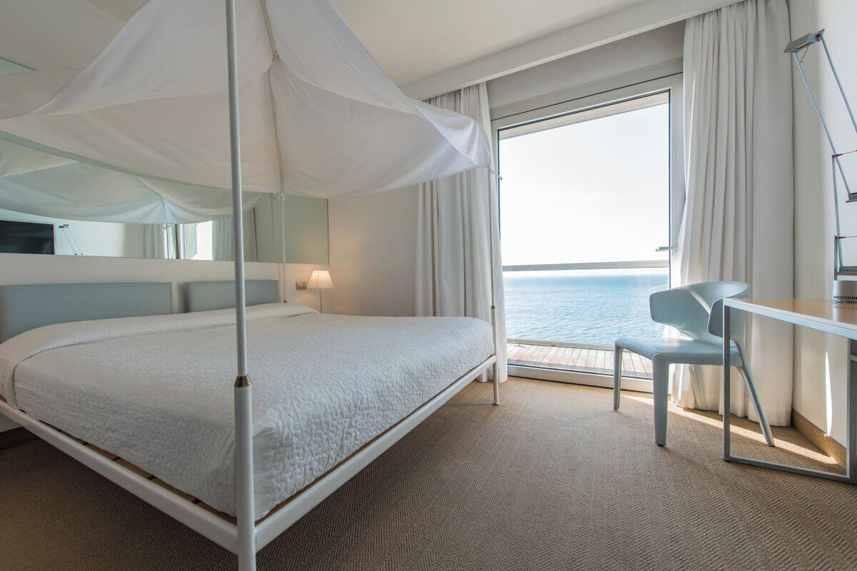 A hotel room with a bed, a desk, a chair, a large window with a view of the sea, and a canopy bed.