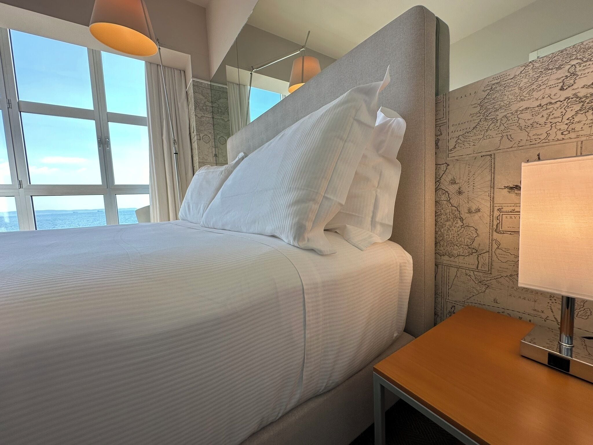 Een hotelkamer met een groot bed, een houten nachtkastje, een kaart aan de muur en uitzicht op zee.