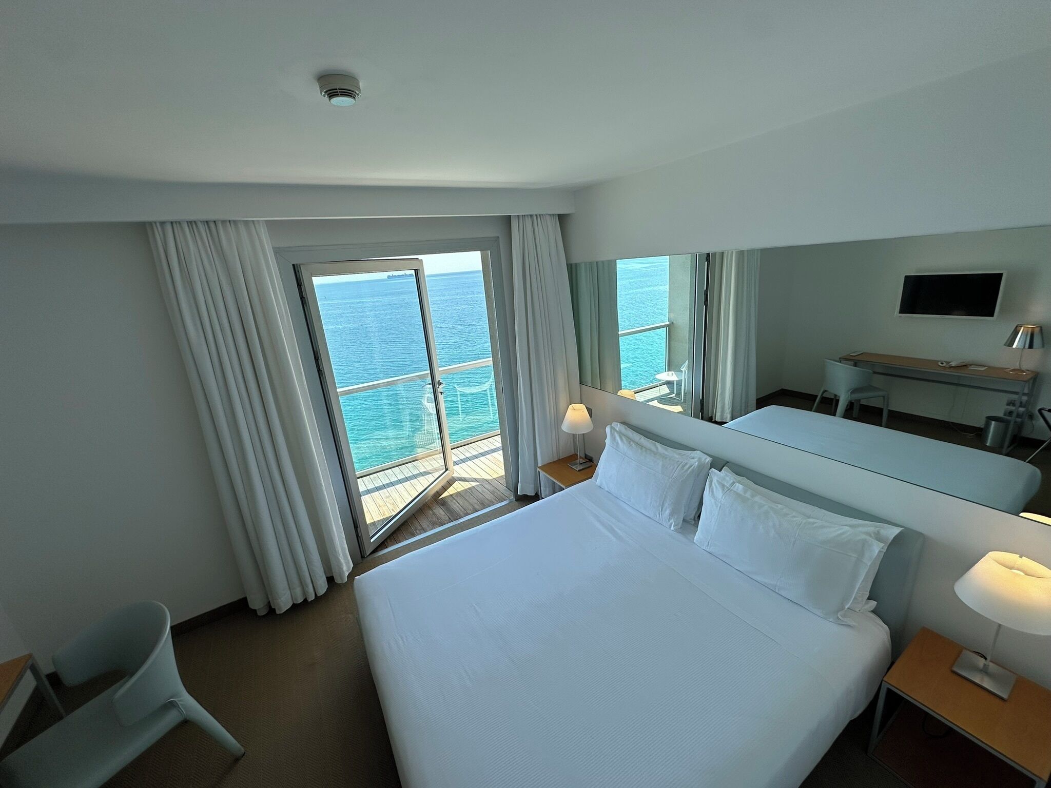 Habitación de hotel con una cama grande, un escritorio, una silla, un televisor y vistas al océano a través de una puerta corrediza.