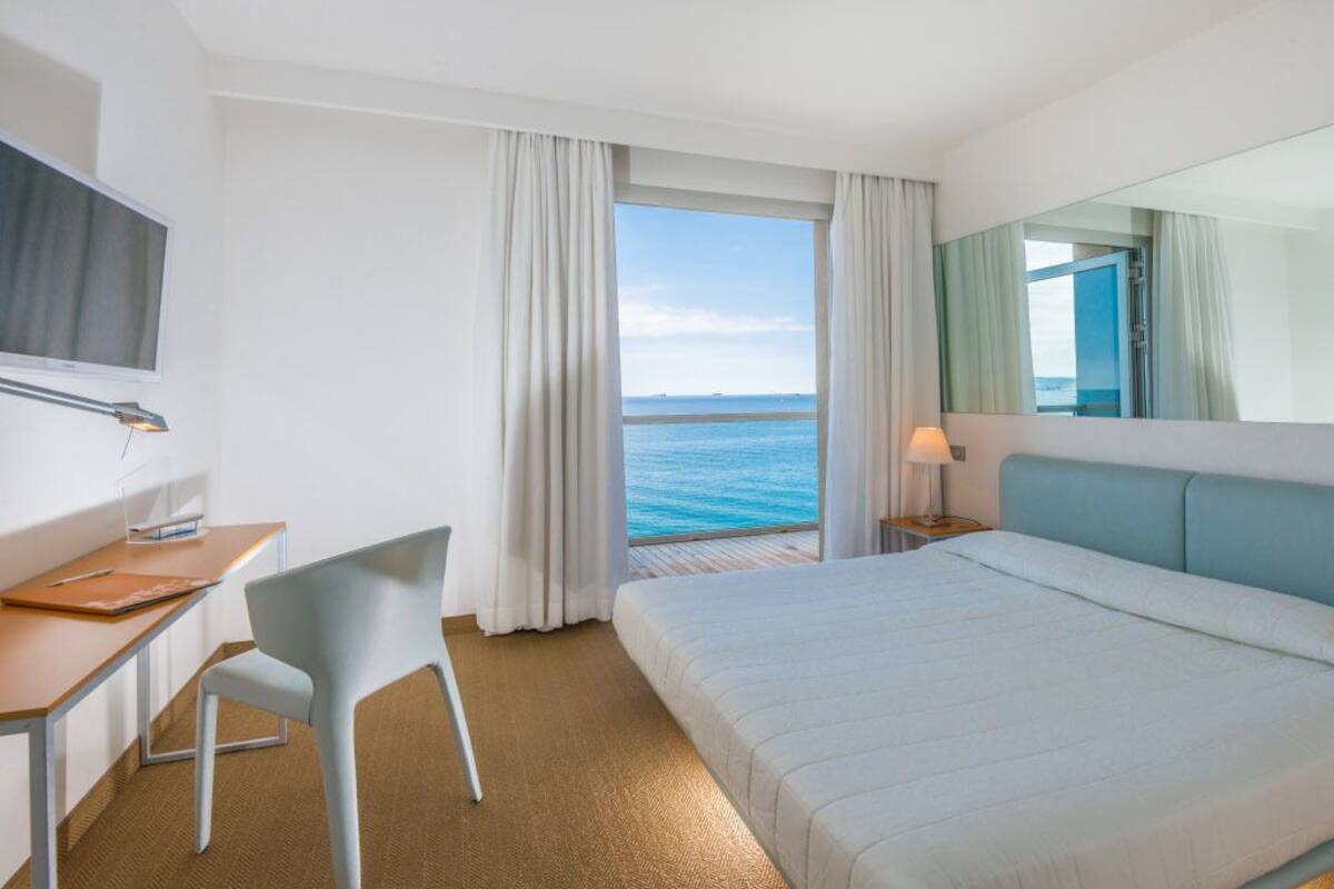 A hotel room with a bed, a desk, a chair, a TV, and a view of the sea.