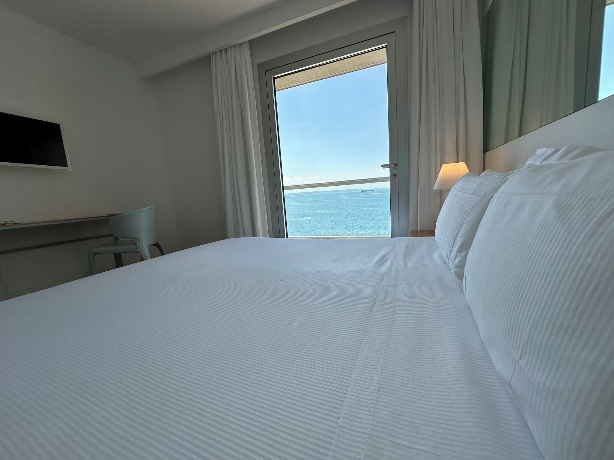 A hotel room with a bed, a desk, a chair, a TV mounted on the wall, and a view of the sea through an open door.