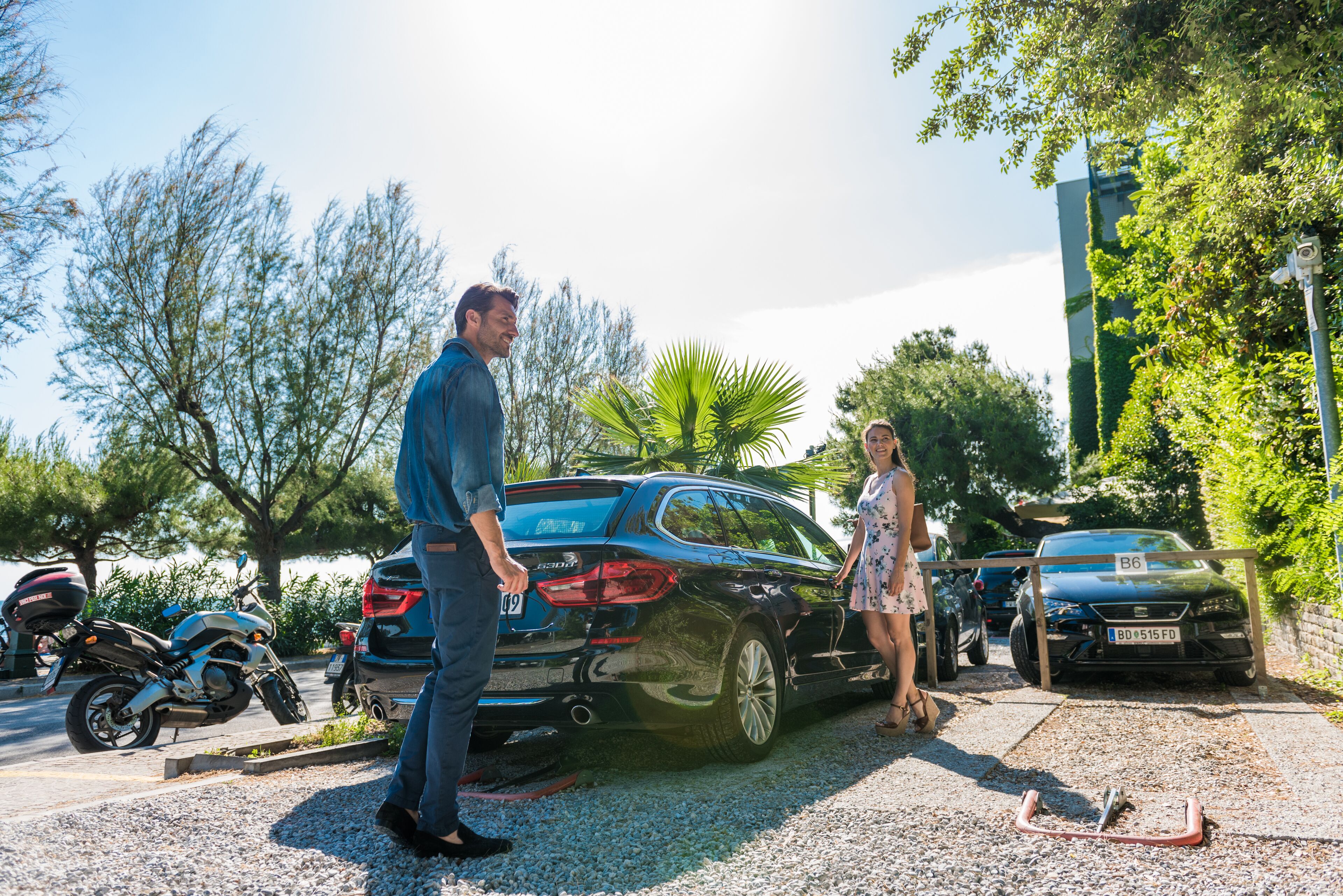 A parking area with a black car, a motorcycle, and a woman standing by the car.