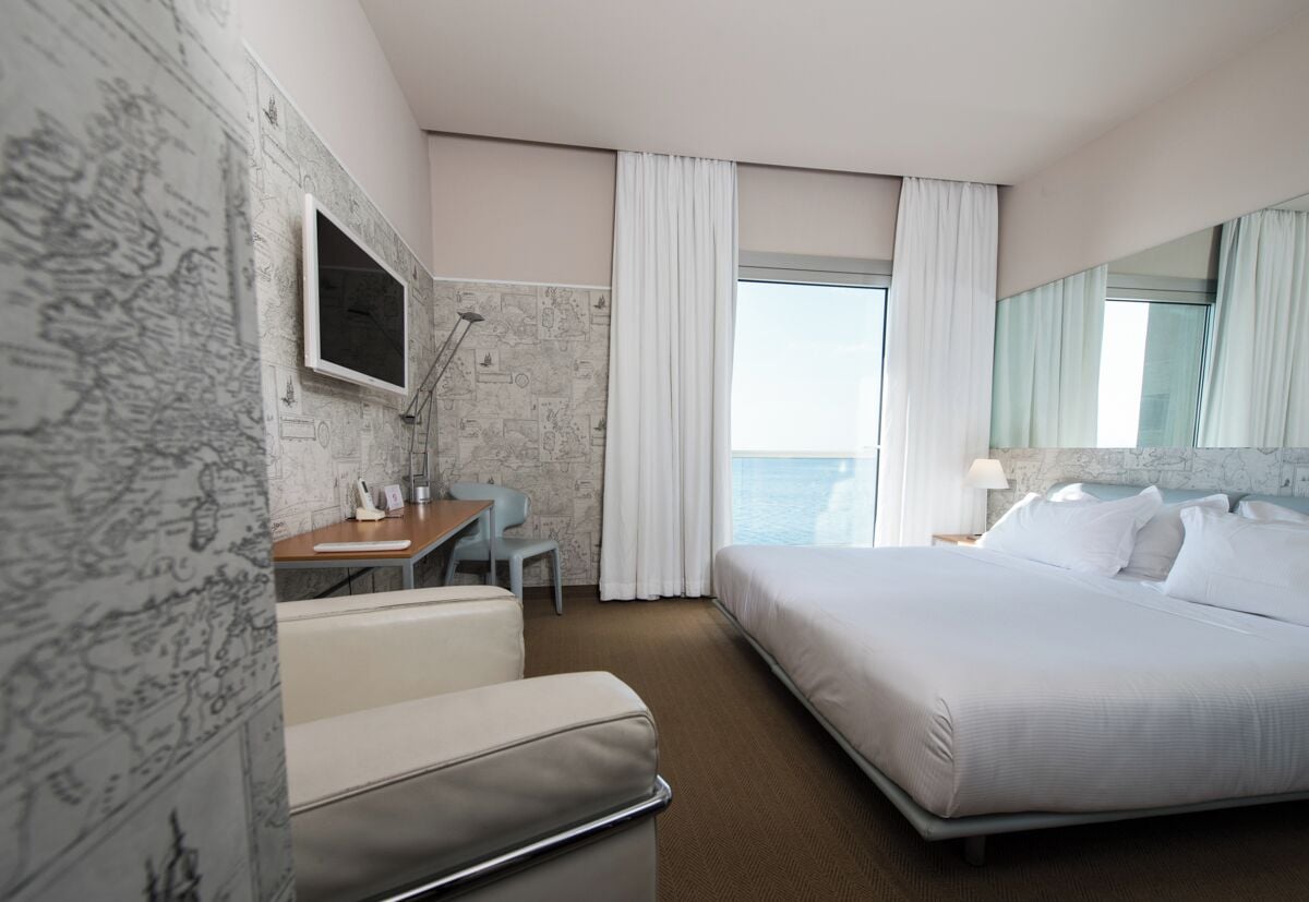 A hotel room with a bed, a desk, a chair, a TV, and a view of the sea.